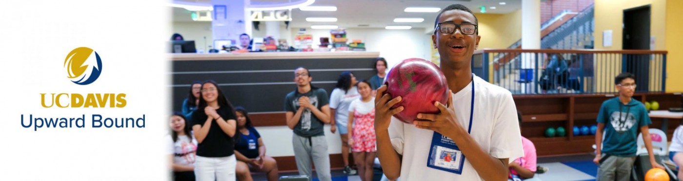 A bowl lifts a bowling ball in a bowling alley with students watching from behind him
